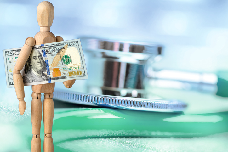 Wooden mannequin holding a US $100 dollar bill on a defocused stethoscope background.