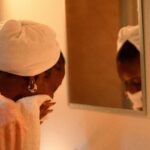 Woman with a towel wrapped around her hair drying her face in front of a mirror